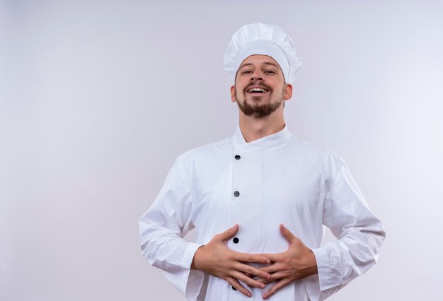 Erfreulicher professioneller männlicher Kochkoch in der weißen Uniform und im Kochhut, der seine Hände auf seinem Bauch hält, der über weißem Hintergrund steht