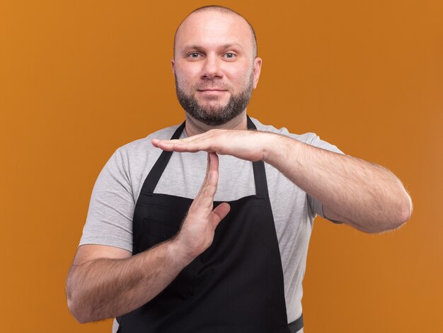 Erfreulicher slawischer männlicher Friseur mittleren Alters in Uniform mit Timeout-Geste isoliert auf orangefarbener Wand
