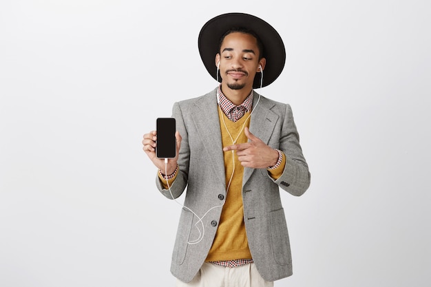 Erfreut attraktiver afroamerikanischer Mann, der Musik in Kopfhörern und Zeigefinger-Smartphonebildschirm hört und Anwendung zeigt