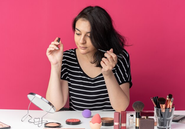 Erfreut Blick auf den Spiegel junges schönes Mädchen sitzt am Tisch mit Make-up-Tools, die Eyeliner isoliert auf rosa Wand halten