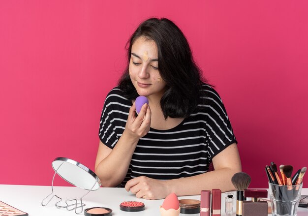 Erfreut Blick auf den Spiegel junges schönes Mädchen sitzt am Tisch mit Make-up-Tools, die Ton-up-Creme mit Schwamm auf rosa Wand auftragen