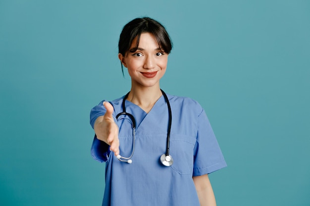 Erfreut, die Hand in die Kamera zu halten, junge Ärztin, die einheitliches Fith-Stethoskop trägt, isoliert auf blauem Hintergrund