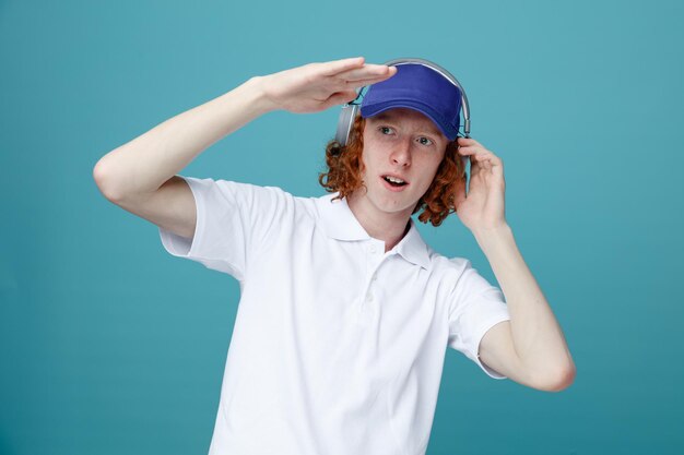 Erfreut erhobene Hand junger gutaussehender Kerl in Mütze mit Kopfhörern isoliert auf blauem Hintergrund