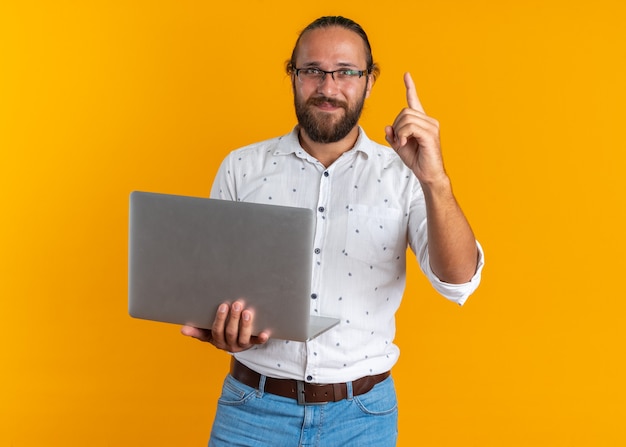 Erfreut erwachsener, gutaussehender Mann mit Brille, der einen Laptop hält und die Kamera anschaut, die isoliert auf einer orangefarbenen Wand nach oben zeigt?