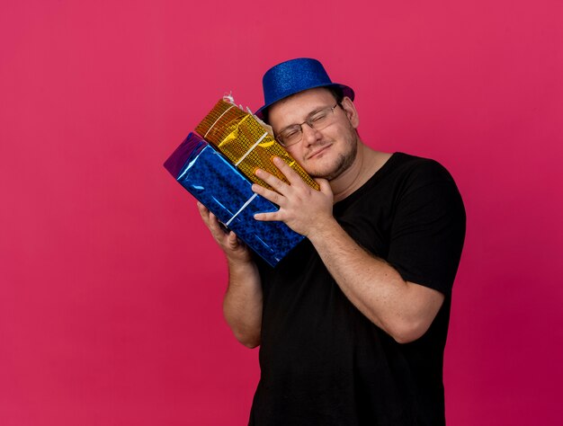Erfreut erwachsener slawischer Mann in optischer Brille mit blauem Partyhut hält und steckt Kopf auf Geschenkboxen