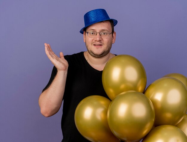 Erfreut erwachsener slawischer Mann in optischer Brille mit blauem Partyhut steht mit erhobener Hand neben Heliumballons