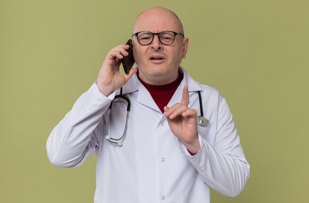 Erfreut erwachsener slawischer Mann mit Brille in Arztuniform mit Stethoskop, der am Telefon spricht und nach oben zeigt