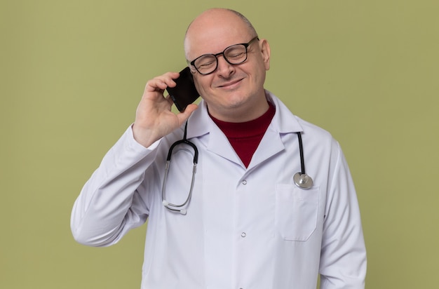 Erfreut erwachsener slawischer Mann mit optischer Brille in Arztuniform mit Stethoskop am Telefon