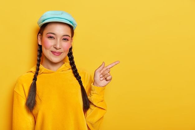 Erfreut fröhliches koreanisches weibliches Model zeigt beiseite, zeigt kühle Leerstelle, trägt Make-up, hat zwei Plaids, trägt Mütze und gelbes Sweatshirt