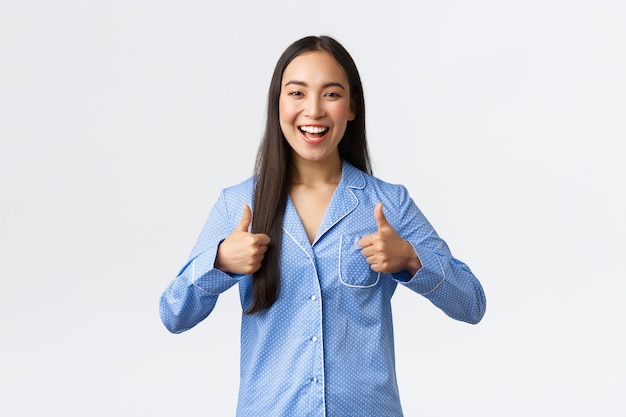 Erfreut glückliches asiatisches Mädchen im blauen Pyjama, das lächelt und Daumen hoch zur Unterstützung zeigt, wie ein tolles Produkt, eine Promo empfehlen, sich über ein großartiges Ergebnis freuen, sagen, gut gespielt oder gute Arbeit?