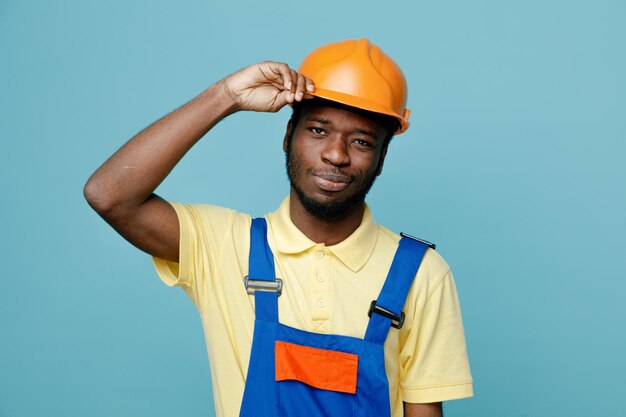 erfreut, Hand auf den Kopf zu legen junger afrikanisch-amerikanischer Baumeister in Uniform isoliert auf blauem Hintergrund