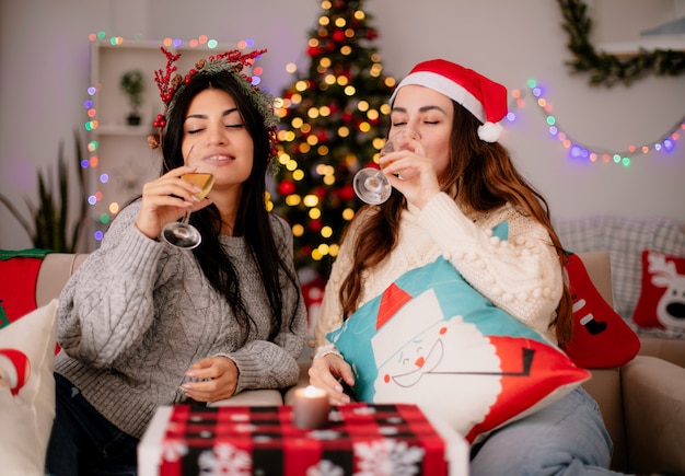 Erfreut hübsche junge Mädchen mit Weihnachtsmütze trinken Gläser Champagner, die auf Sesseln sitzen und die Weihnachtszeit zu Hause genießen