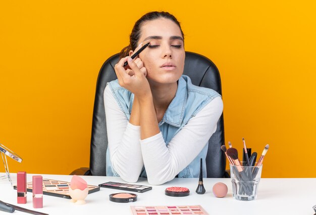 Erfreut hübsche kaukasische Frau, die am Tisch mit Make-up-Tools sitzt, die Eyeliner auftragen