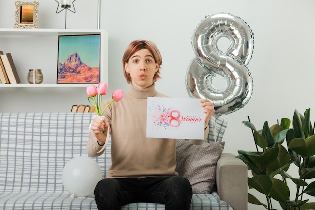 Erfreut hübscher Kerl am glücklichen Frauentag, der Blumen mit Grußkarte auf dem Sofa im Wohnzimmer hält