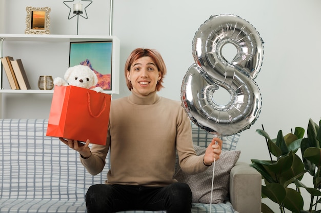 Erfreut hübscher Kerl am glücklichen Frauentag, der den Ballon Nummer acht mit der Geschenktüte auf dem Sofa im Wohnzimmer hält