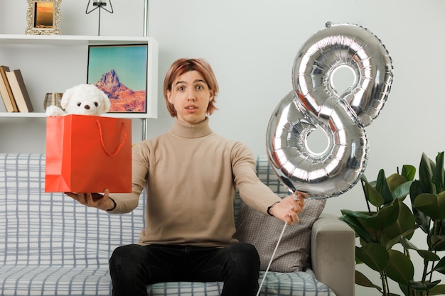 Erfreut hübscher Kerl am glücklichen Frauentag, der den Ballon Nummer acht mit Geschenktüte hält und auf dem Sofa im Wohnzimmer sitzt