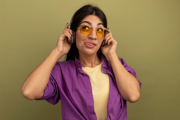 Erfreut hübsches brünettes kaukasisches Mädchen in Sonnenbrille mit Kopfhörern steckt Zunge heraus und schaut zur Seite auf Olivgrün