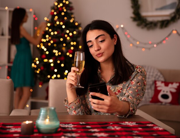 Erfreut hübsches junges Mädchen schmückt Weihnachtsbaum und ihre Freundin hält ein Glas Champagner und macht Selfie am Tisch sitzen und die Weihnachtszeit zu Hause genießen enjoying