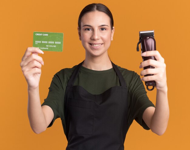 Erfreut junge brünette Friseurin in Uniform mit Haarschneidemaschine und Kreditkarte and