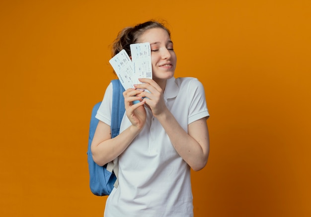 Erfreut junge hübsche Studentin, die Rückentasche hält, die Flugtickets mit geschlossenen Augen lokalisiert auf orange Hintergrund mit Kopienraum hält