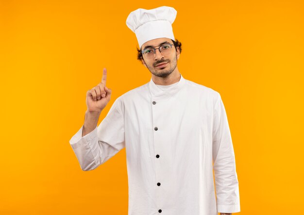 erfreut junge männliche Köchin in Kochuniform und Brille zeigt nach oben