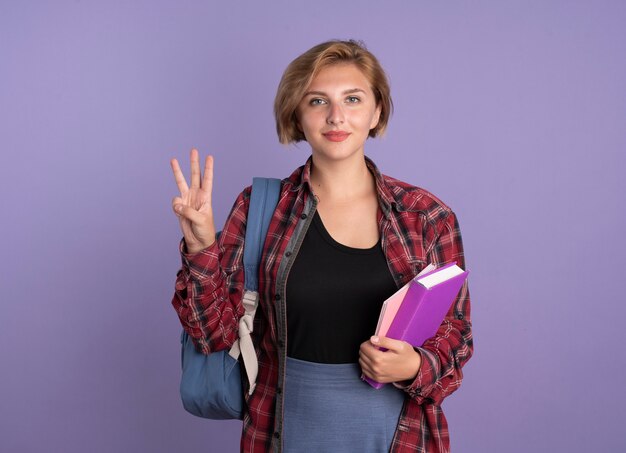 Erfreut junge slawische Studentin mit Rucksack hält Buch und Notizbuch Gesten drei mit den Fingern