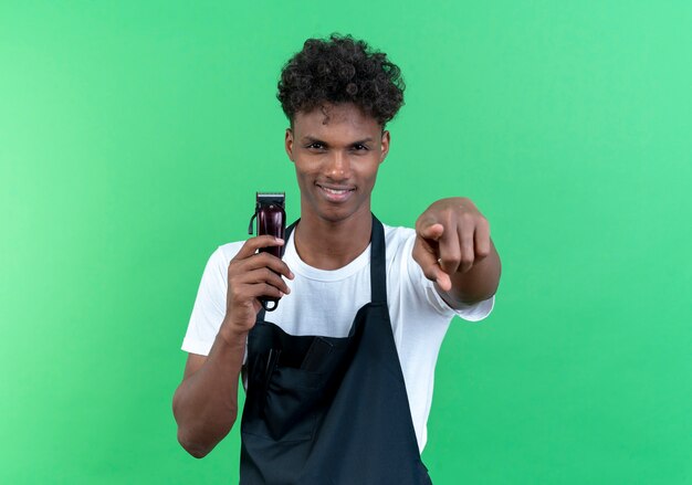 Erfreut junger afroamerikanischer männlicher Friseur, der eine Uniform trägt, die Haarschneidemaschinen hält und Ihnen Geste zeigt