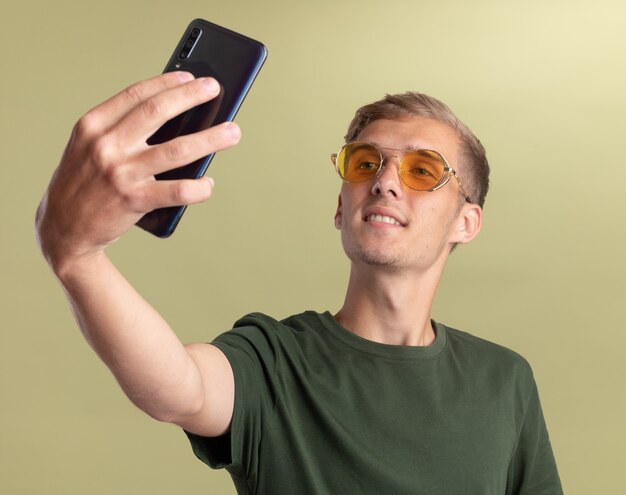Erfreut junger gutaussehender Kerl mit grünem Hemd mit Brille macht ein Selfie isoliert auf olivgrüner Wand