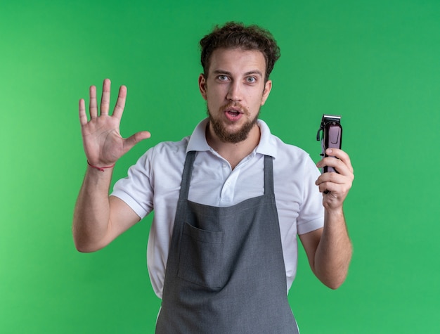 Erfreut junger männlicher Friseur, der eine Uniform trägt, die Haarschneidemaschinen hält und die Stoppgeste einzeln auf grüner Wand zeigt