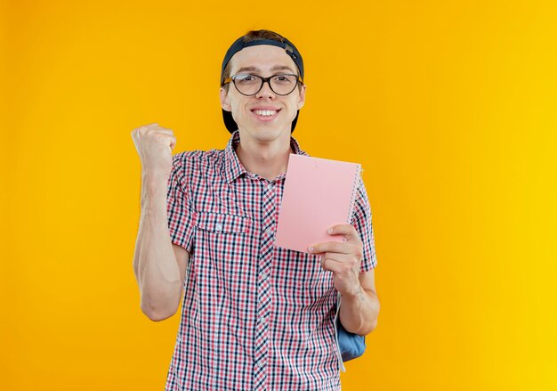Erfreut junger Studentenjunge, der eine Tasche und eine Brille und eine Mütze trägt, die ein Notizbuch hält und eine Ja-Geste zeigt