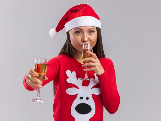 Erfreut junges asiatisches Mädchen mit Weihnachtsmütze mit Pullover mit Glas Champagner und Getränken isoliert auf weißem Hintergrund