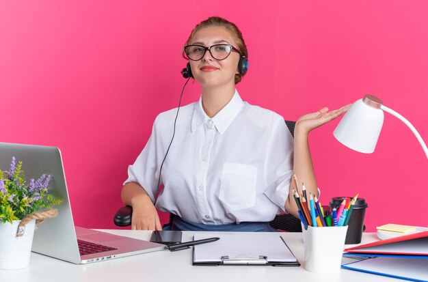 Erfreut junges blondes Callcenter-Mädchen mit Headset und Brille, das am Schreibtisch mit Arbeitswerkzeugen sitzt und leere Hand zeigt, die auf die Kamera isoliert auf rosa Wand blickt