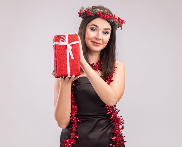 Erfreut junges hübsches kaukasisches Mädchen mit Weihnachtskopfkranz und Lametta-Girlande um den Hals, das Geschenkpaket hält und die Kamera einzeln auf weißem Hintergrund mit Kopierraum betrachtet