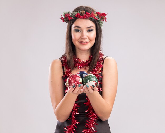 Erfreut junges hübsches kaukasisches Mädchen mit Weihnachtskopfkranz und Lametta-Girlande um den Hals, das Weihnachtskugeln mit Blick auf die Kamera isoliert auf weißem Hintergrund mit Kopienraum hält