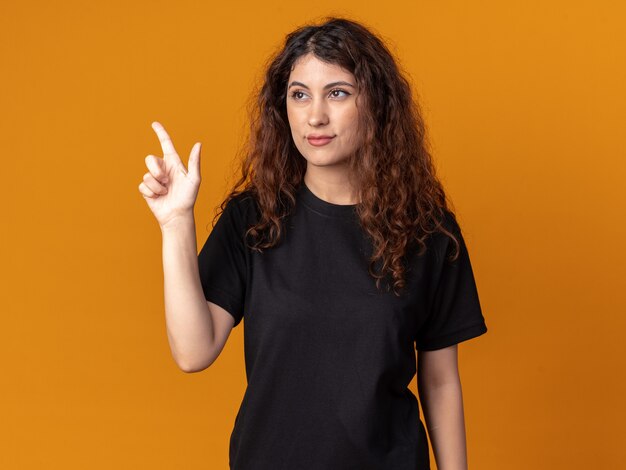 Erfreut junges hübsches Mädchen, das auf die Seite schaut, die nach oben zeigt, isoliert auf der orangefarbenen Wand?