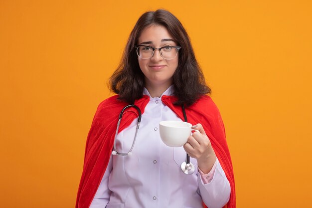 Erfreut junges kaukasisches Superheldenmädchen in Arztuniform und Stethoskop mit Brille, die eine Tasse Tee hält