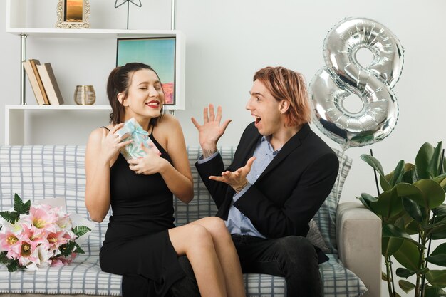 Erfreut junges Paar am glücklichen Frauentag Mädchen, das den anwesenden Kerl hält, der die Hände auf dem Sofa im Wohnzimmer ausbreitet