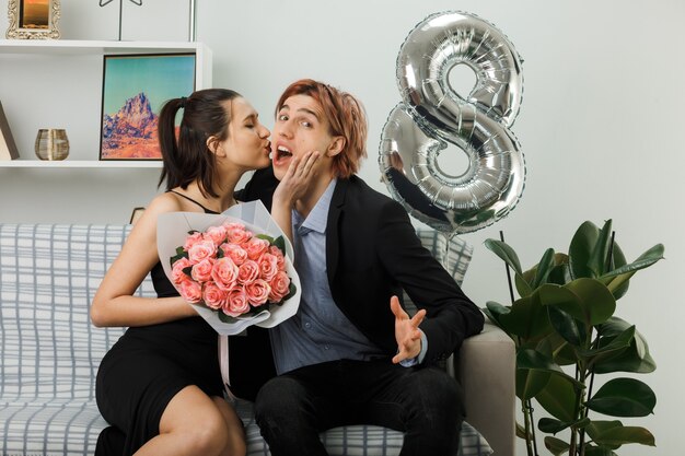 Erfreut junges Paar am glücklichen Frauentag Mädchen mit Blumenstrauß küssenden Kerl sitzt auf dem Sofa im Wohnzimmer