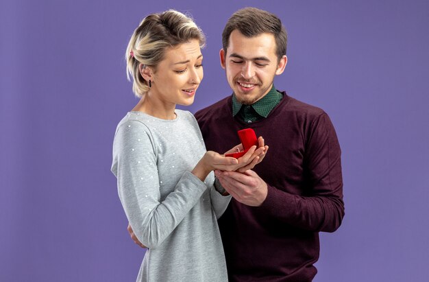 Erfreut junges Paar am Valentinstag mit Blick auf Ehering in Mädchenhänden isoliert auf blauem Hintergrund