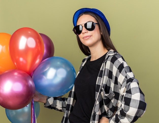 Erfreut junges schönes Mädchen mit blauem Hut mit Brille, die Luftballons hält, die Hand auf die Hüfte legt