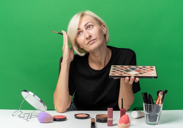 Erfreut junges schönes Mädchen sitzt am Tisch mit Make-up-Tools, die Lidschatten-Palette mit Make-up-Pinsel auf grüner Wand halten