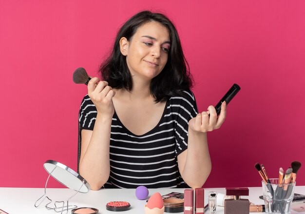 Erfreut junges schönes Mädchen sitzt am Tisch mit Make-up-Tools, die Puderpinsel mit Mascara isoliert auf rosa Wand halten