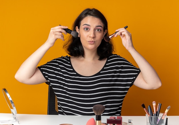 Erfreut junges schönes Mädchen sitzt am Tisch mit Make-up-Tools, die Puderpinsel um das Gesicht halten, isoliert auf oranger Wand