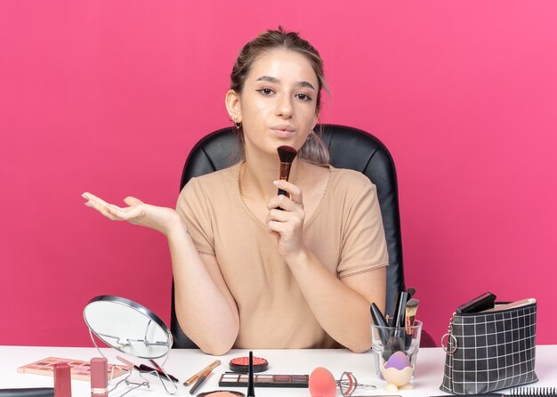 Erfreut junges schönes Mädchen sitzt am Tisch mit Make-up-Tools, die Pulverpinsel auf rosa Wand isoliert halten und blasen