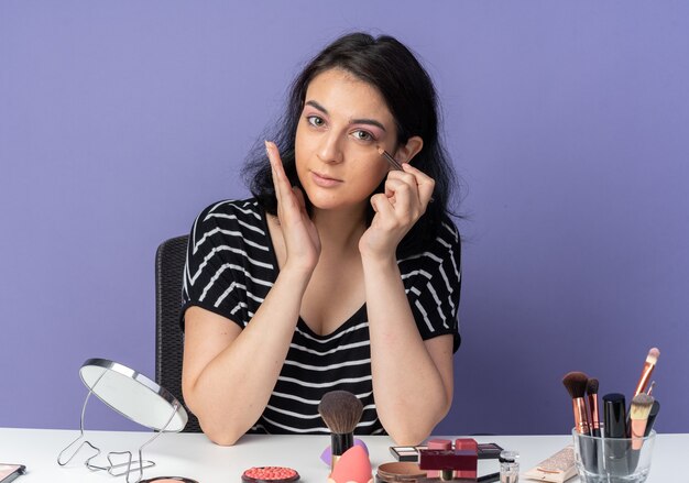 Erfreut junges schönes Mädchen sitzt am Tisch mit Make-up-Tools ziehen Pfeil mit Eyeliner isoliert auf blauer Wand