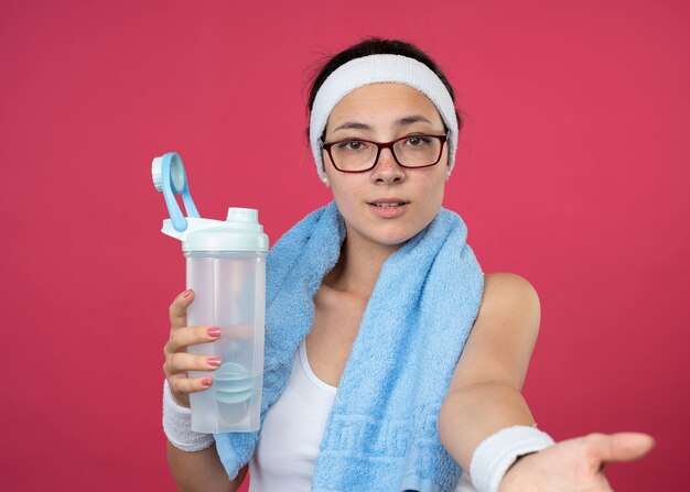 Erfreut junges sportliches Mädchen in optischer Brille mit Handtuch um den Hals, das Stirnband und Armbänder trägt, die eine Wasserflasche halten und die Hand ausstrecken