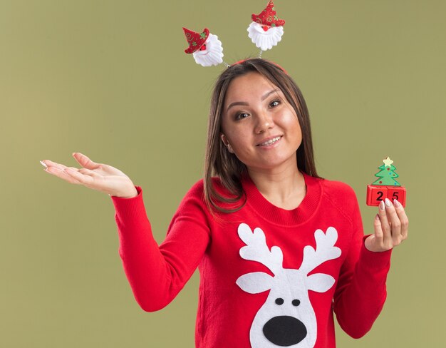 Erfreut kippendes junges asiatisches Mädchen des Kippkopfes, das Weihnachtshaarbügel hält, der Weihnachtsspielzeugpunkte mit Hand an der Seite lokalisiert auf olivgrüner Wand hält