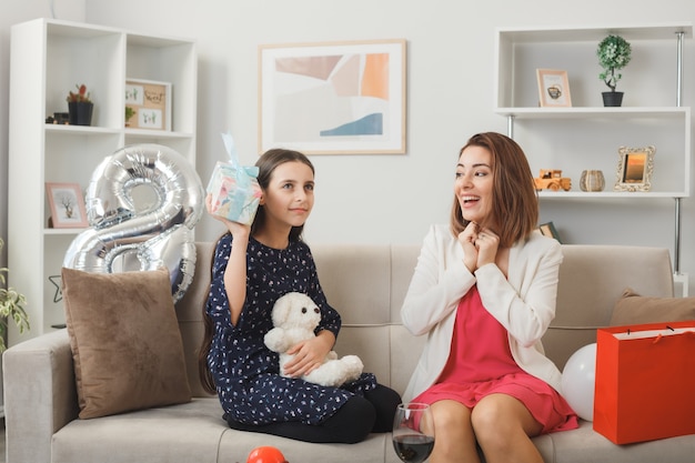 Erfreut kleines Mädchen und Mather mit Geschenk und Teddybär mit Geschenk am glücklichen Frauentag sitzen auf dem Sofa im Wohnzimmer