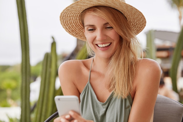 Erfreut lächelnde junge Frau mit fröhlichem Ausdruck trägt Sommerkleidung, freut sich, Nachricht zu erhalten oder liest positive Nachrichten online auf Smartphone, verbunden mit drahtlosem Internet im Straßencafé