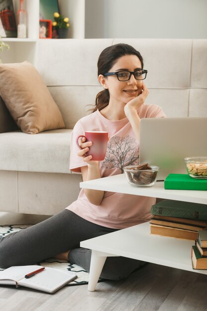 Erfreut mit der Hand unter dem Kinn junges Mädchen, das eine Tasse Tee mit Brille auf dem Boden hinter dem Couchtisch im Wohnzimmer hält